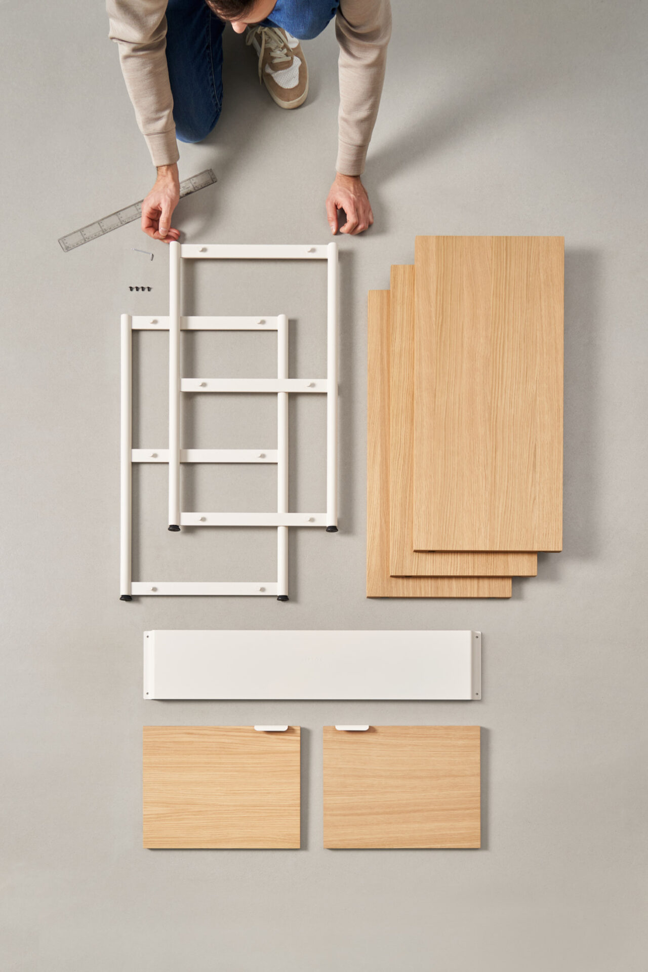 Person assembling Tiptoe furniture components laid out on the floor, including wooden shelves and white metal supports.